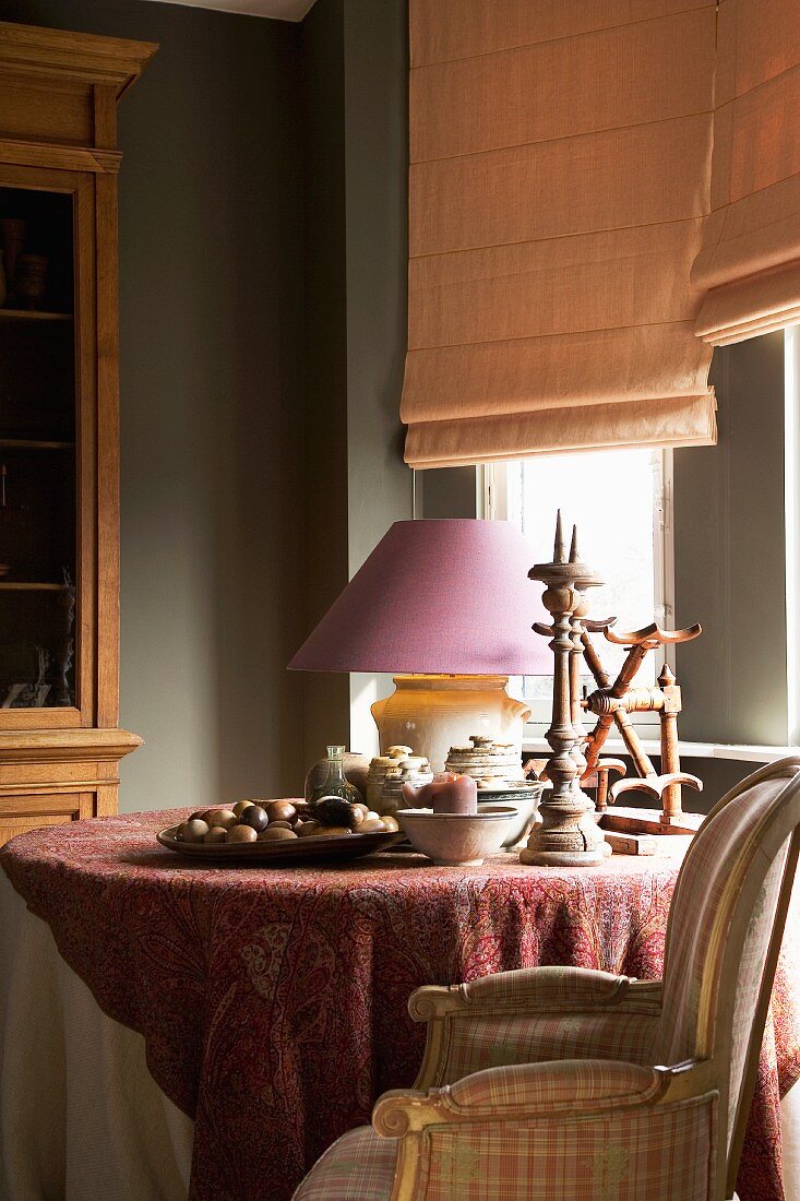 Round table decorated with old wooden candlesticks and table lamp with pastel purple lampshade below window