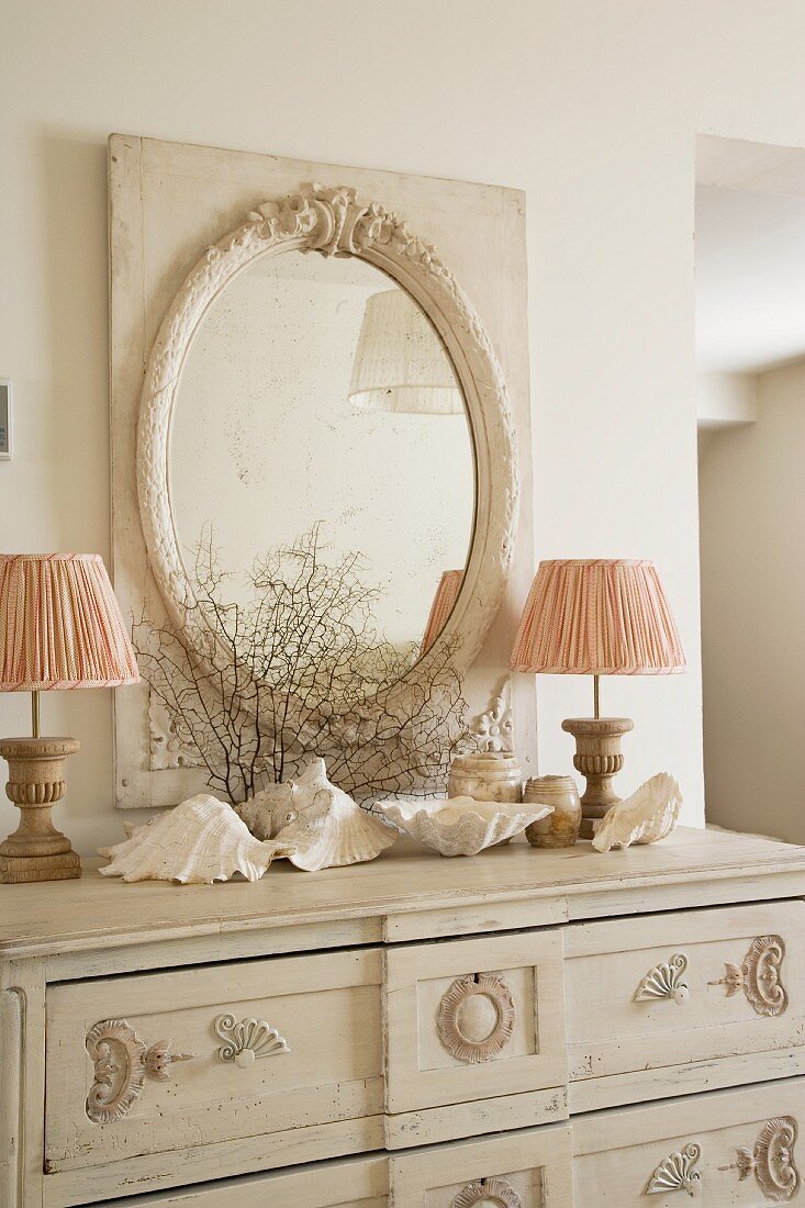 Feminine oval mirror, table lamps and collection of sea shells on antique chest of drawers