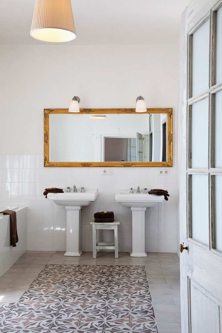 Blick durch offene Badtür auf zwei Standwaschbecken vor Spiegel mit Goldrahmen und Fliesenboden mit Blumenmuster