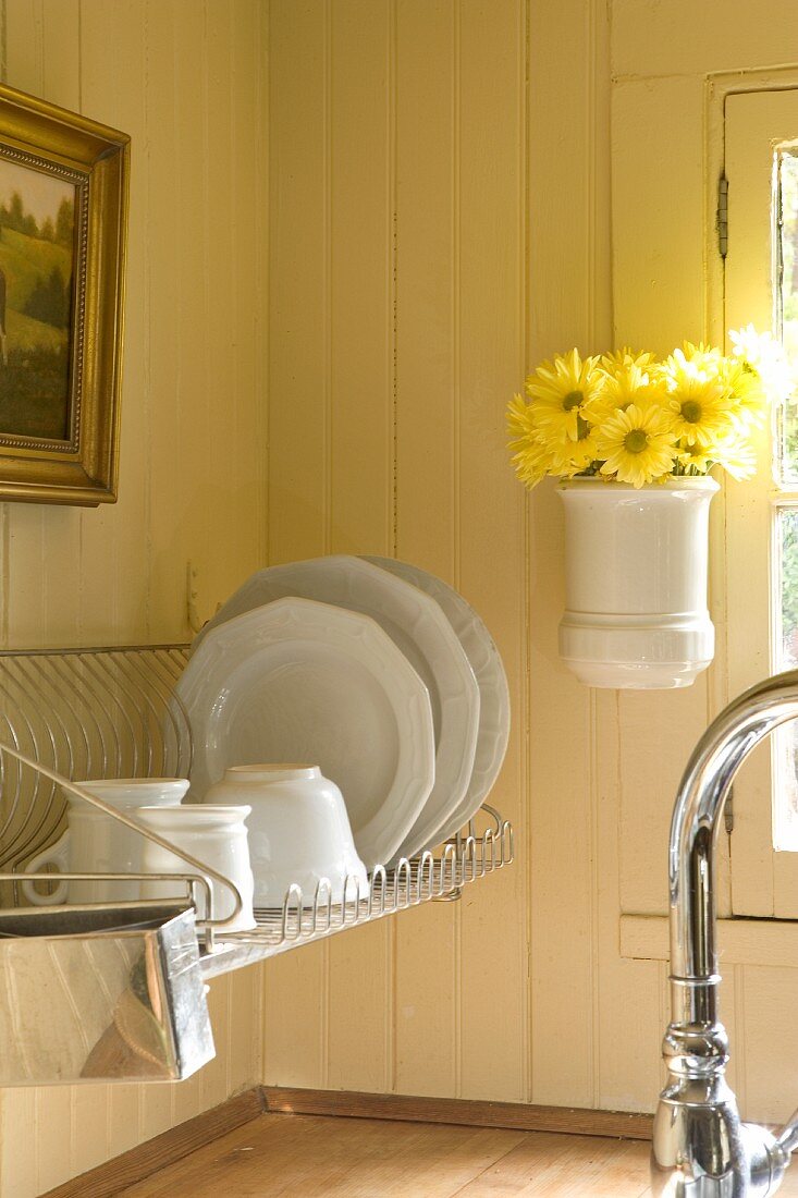 White crockery on draining rack next to vase of yellow flowers hung on wood-clad wall