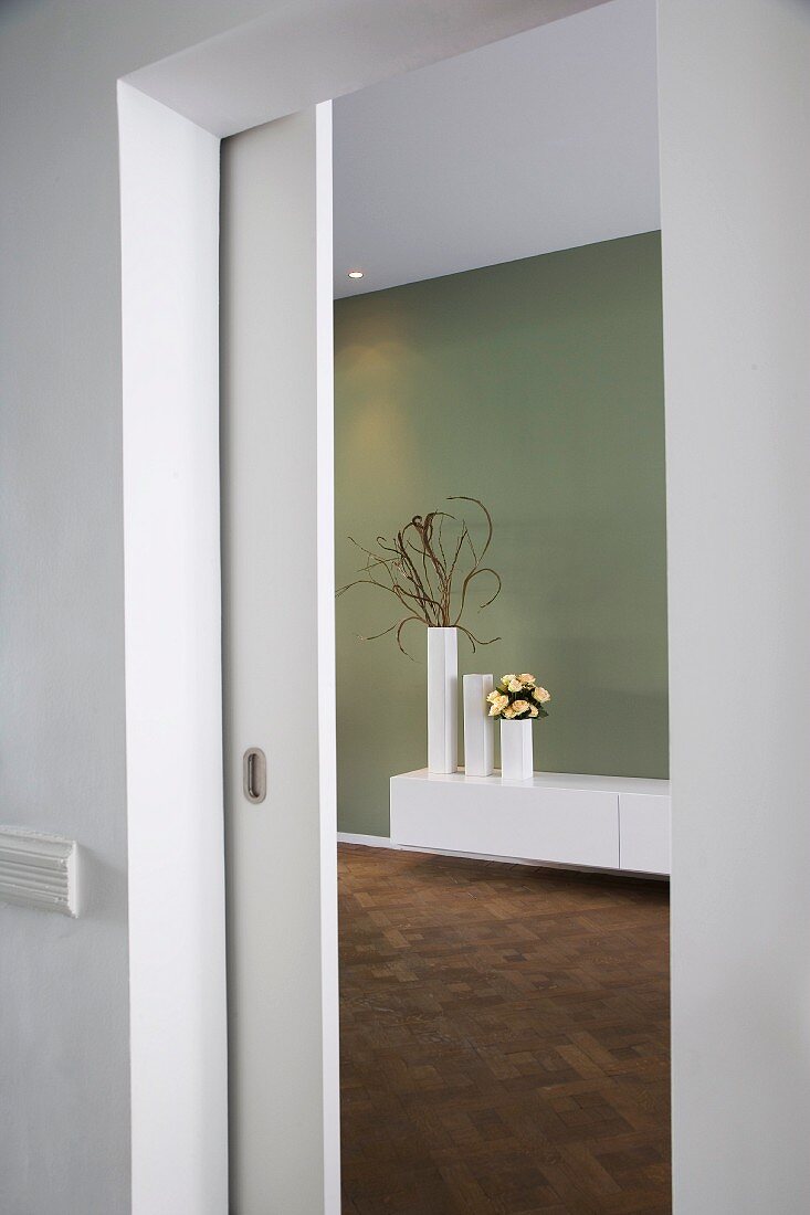 Blick durch offene Schiebetür auf weisses Wandbord mit Vasengruppe vor grau getönter Wand