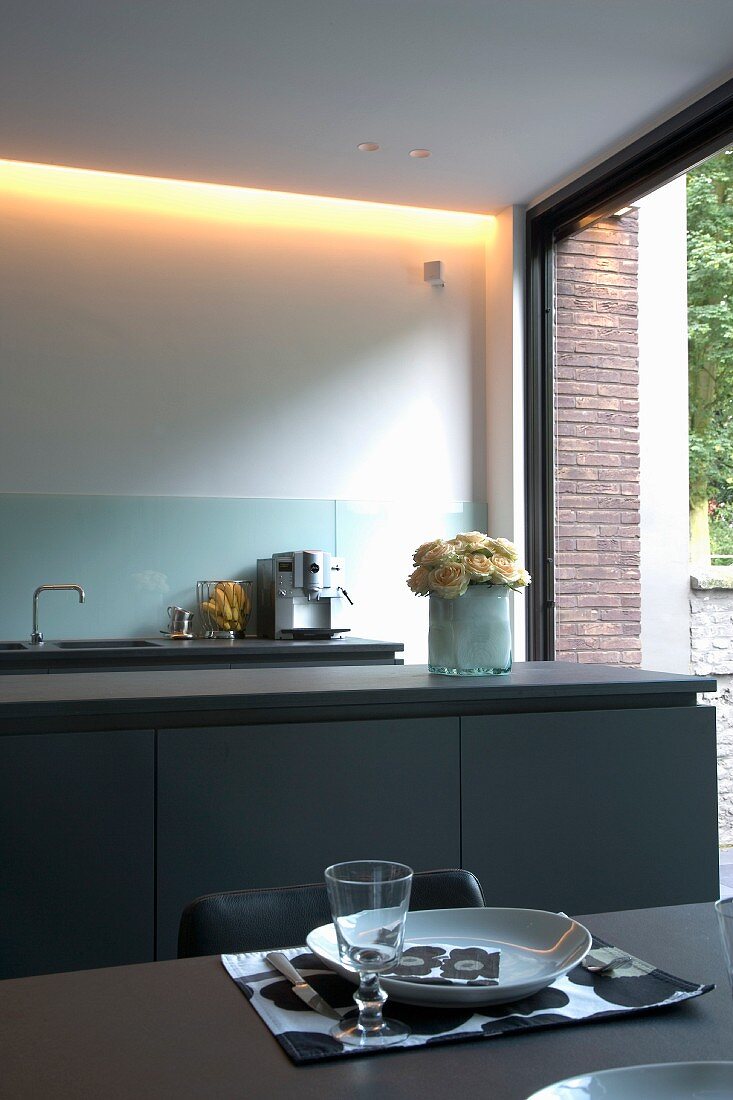 Place setting on dining table in front of kitchen counter with grey base units and indirect lighting next to open terrace door