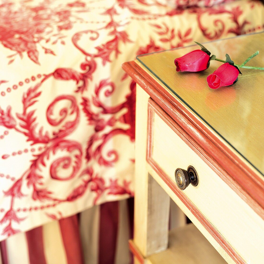 Künstliche Rosen auf Nachttisch mit weiss roter Bemalung neben Bett mit gemusterter Tagesdecke in traditionellem Stil