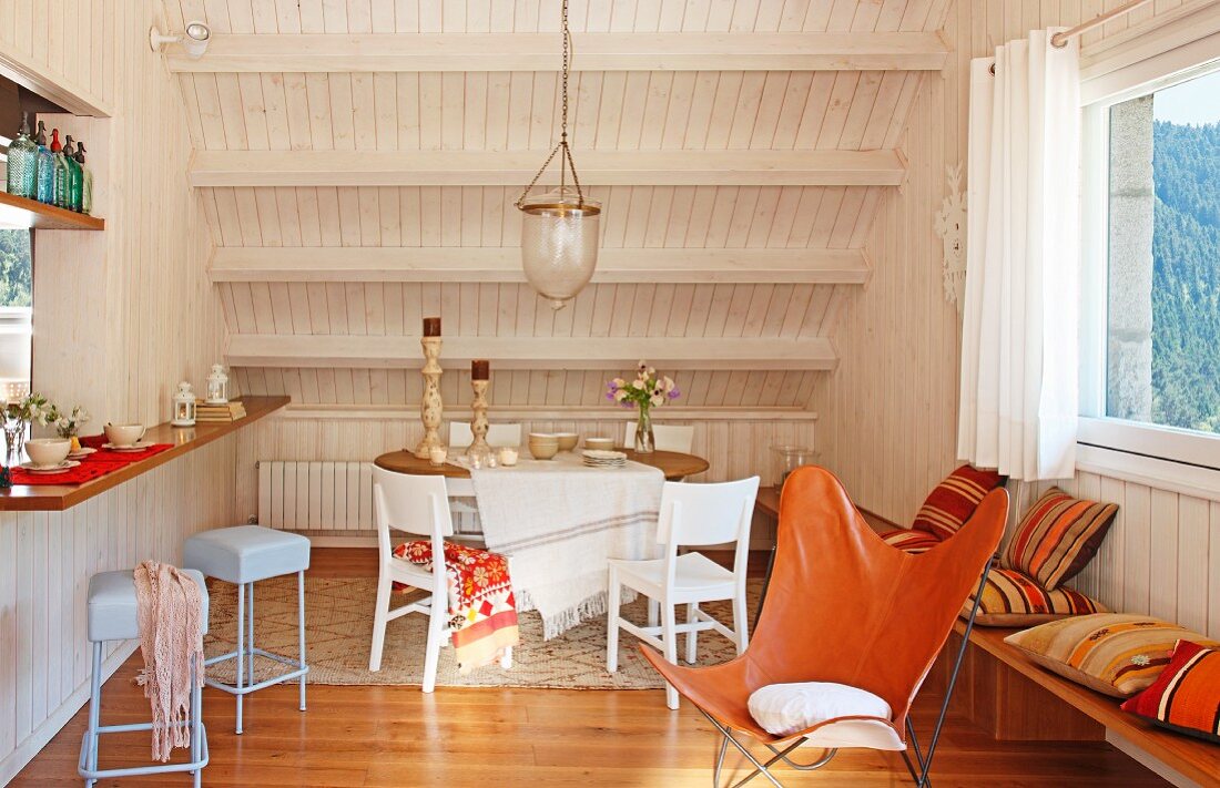 Leather butterfly chair in front of dining area with white chairs below wood-clad sloping ceiling