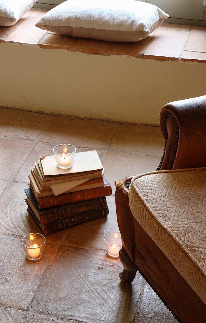 Partially visible leather armchair next to stack of books and lit tealights in front of masonry bench with cushions