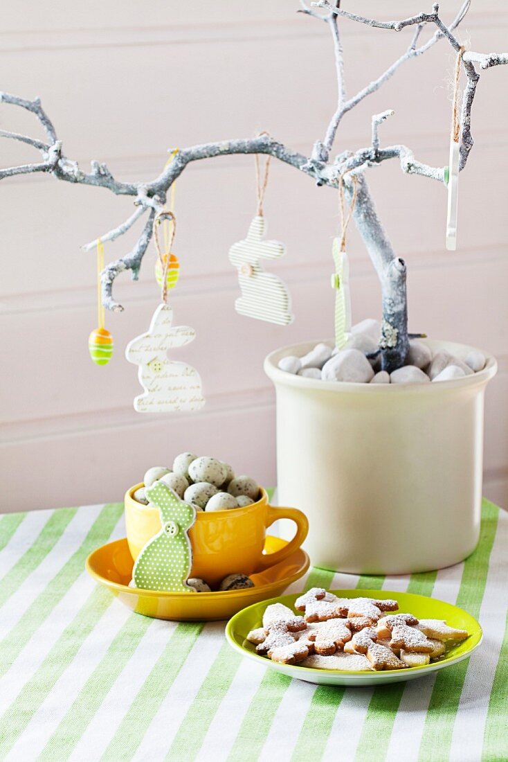 Easter arrangement with rabbits and eggs hanging from branch above cup of quail's eggs and plate of Easter biscuits