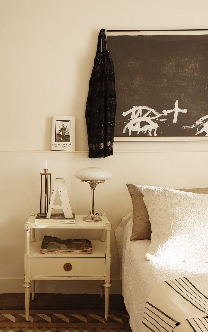 Antique bedside table with retro-style table lamp and dark artwork on wall above bed