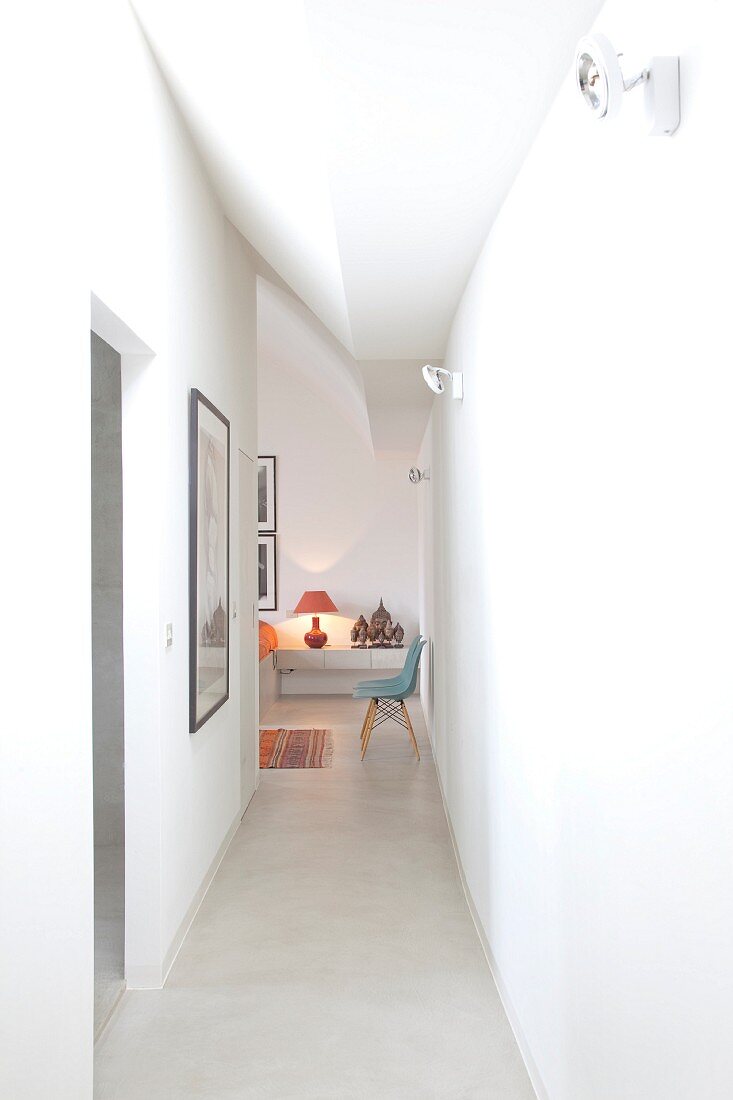 Narrow, white corridor with view of classic 50s chair and lit red table lamp in adjoining room
