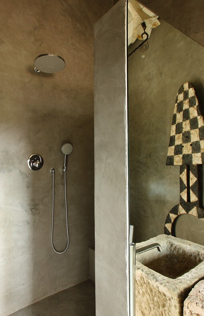 Betonfarben gespachteltes Designerbad mit offener Dusche und Waschtrog aus Naturstein; im Spiegel reflektierte Ethnokunst