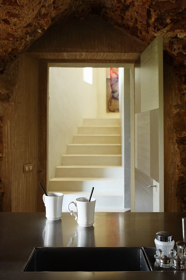 Zwei Kaffeebecher vor dem Unterbaubecken einer Edelstahlarbeitsfläche; im Hintergrund Blick durch offene Tür auf gemauerte Treppe