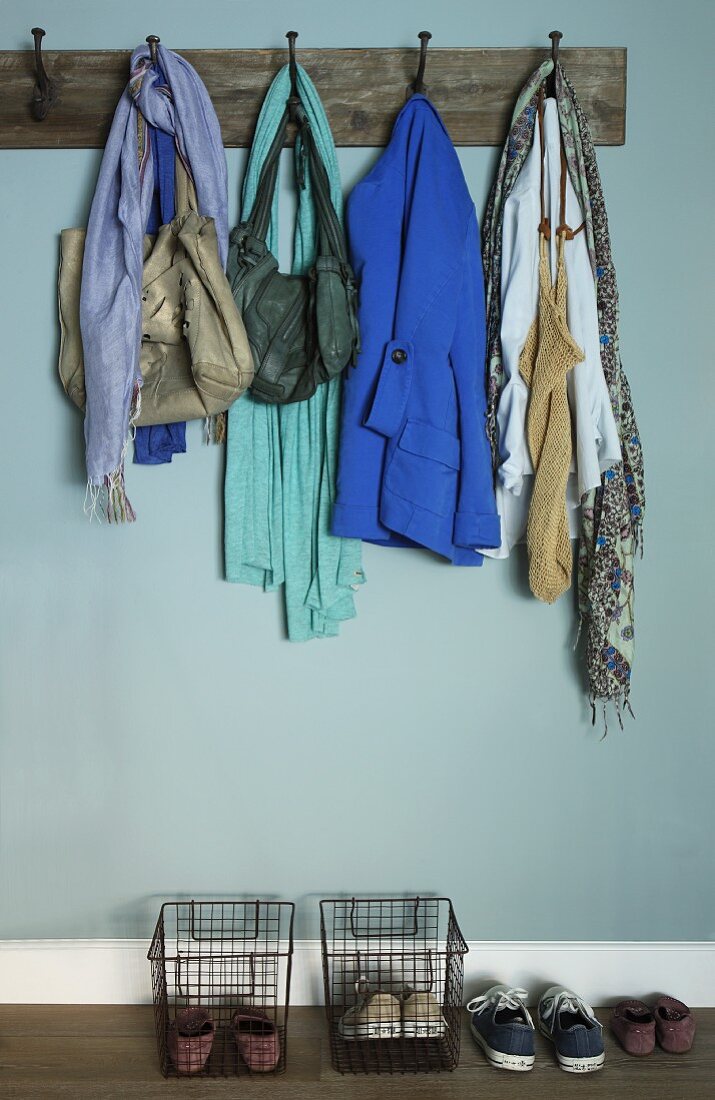 Offene Garderobe - Vintage Wandhakenleiste aus Holz mit Kleidung und Schuhe in Drahtkörben vor hellblau getönter Wand