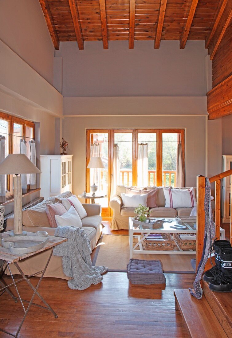 Blick von Treppe in Loungebereich mit heller Sofagarnitur vor Balkonfenster in offenem Wohnraum mit Holzdecke