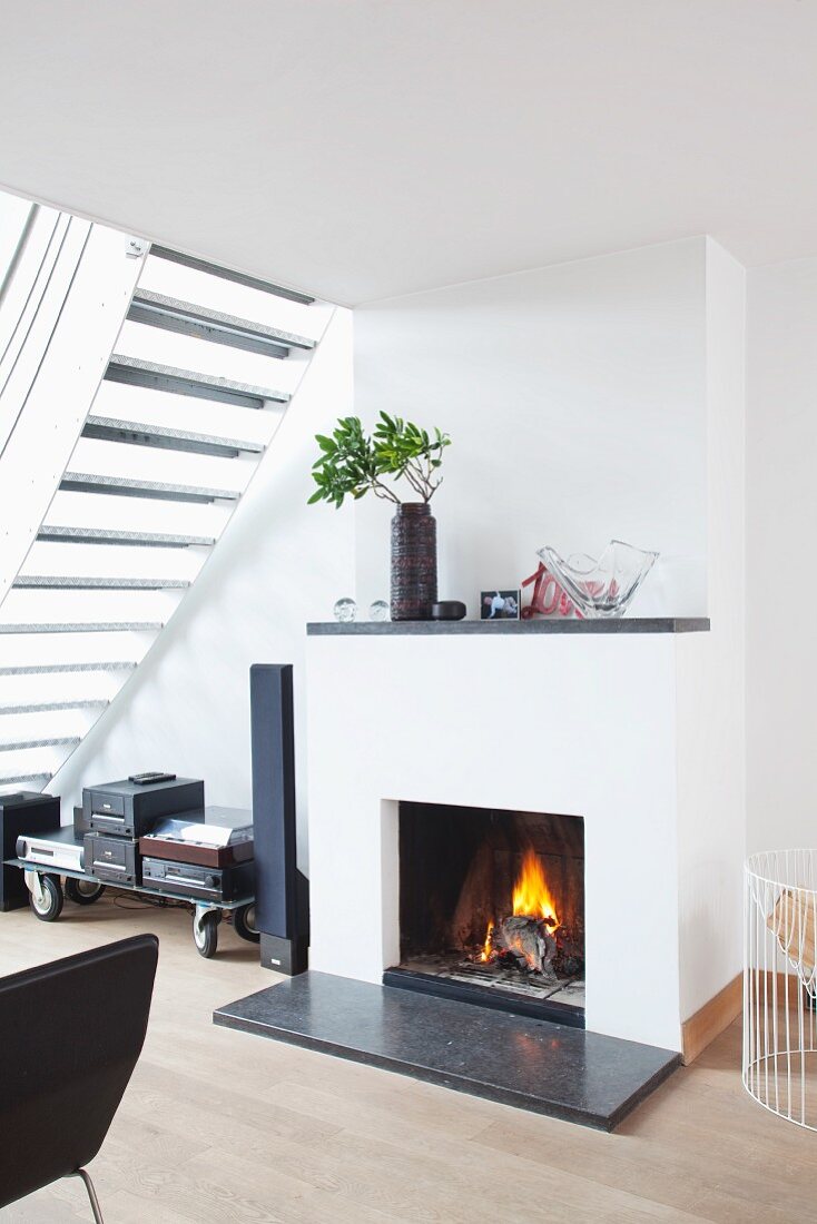 Fire in modern fireplace with dark stone hearth and mantelpiece next to hifi system on trolley under airy staircase