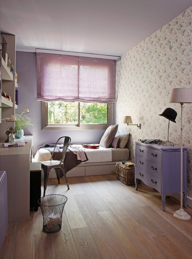 Fliederfarbenes Jugendzimmer - Lila bemalte Kommode im Neo-Barockstil vor Wand mit gemusterter Tapete und Bett am Fenster mit halbgeschlossenem Rollo