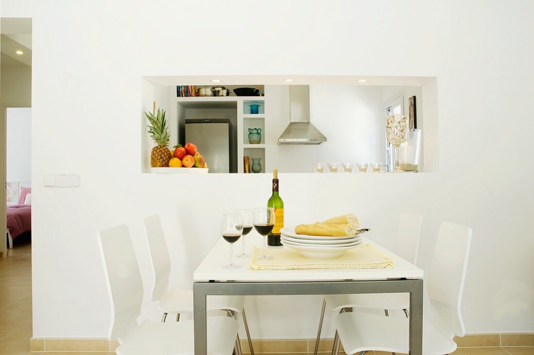 White, modern dining area with red wine and bread on table in front of service hatch to kitchen