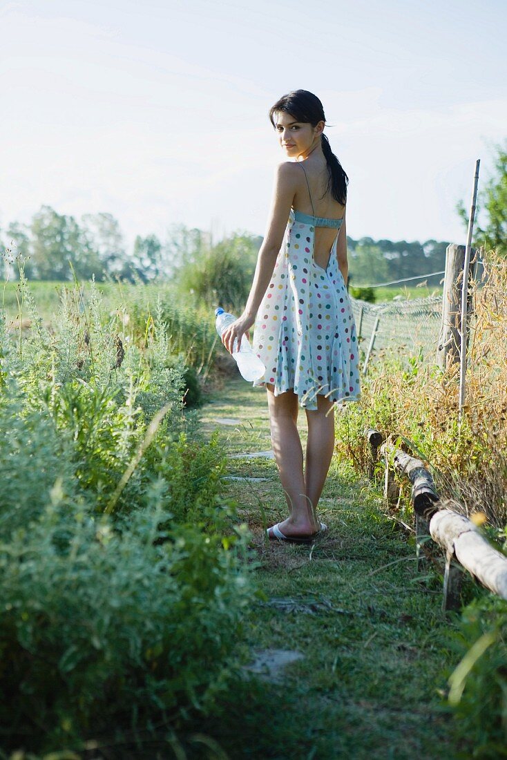 Young woman standing in garden with bottle of water, looking over shoulder at camera