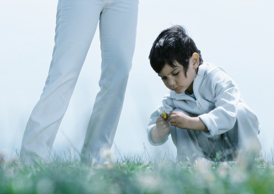 Little boy squatting down on grass looking at flower next to woman's legs