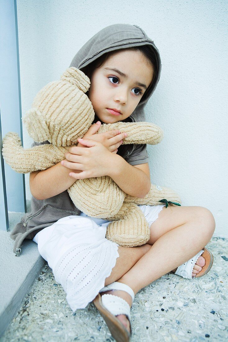 Kind mit Teddybär im Arm sitzt vor der Hauswand