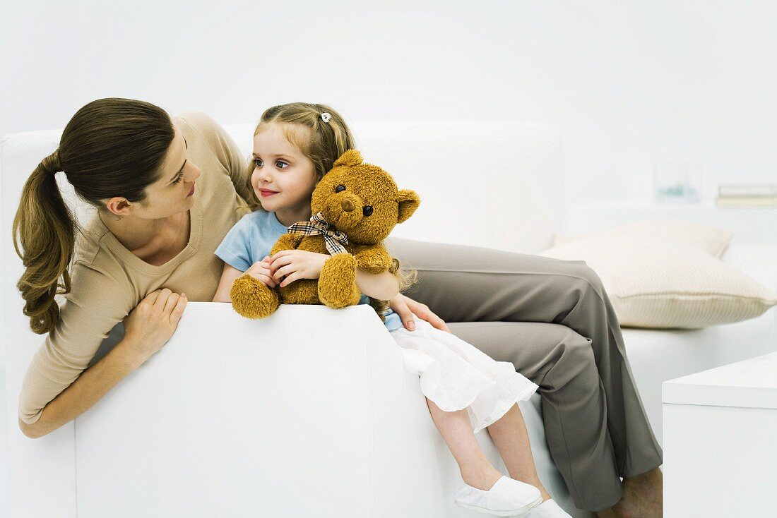 Mother and daughter sitting on couch, smiling at each other, girl holding teddy bear