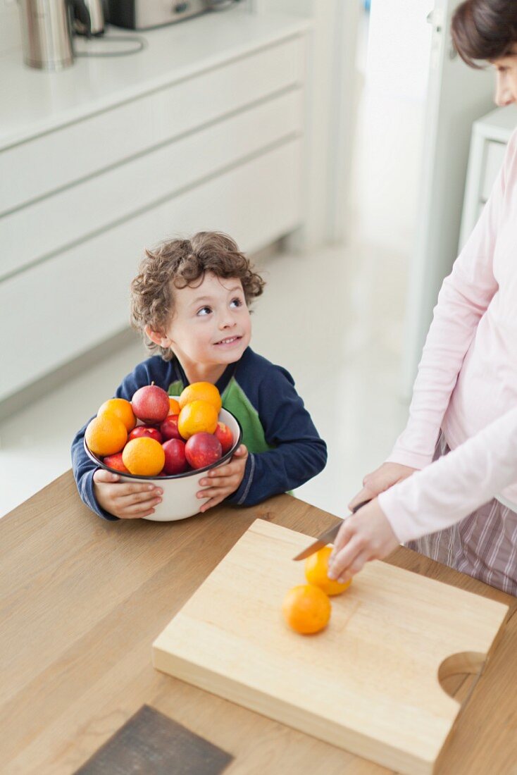 A mother cutting oranges and a boy holding a bowl of fruit