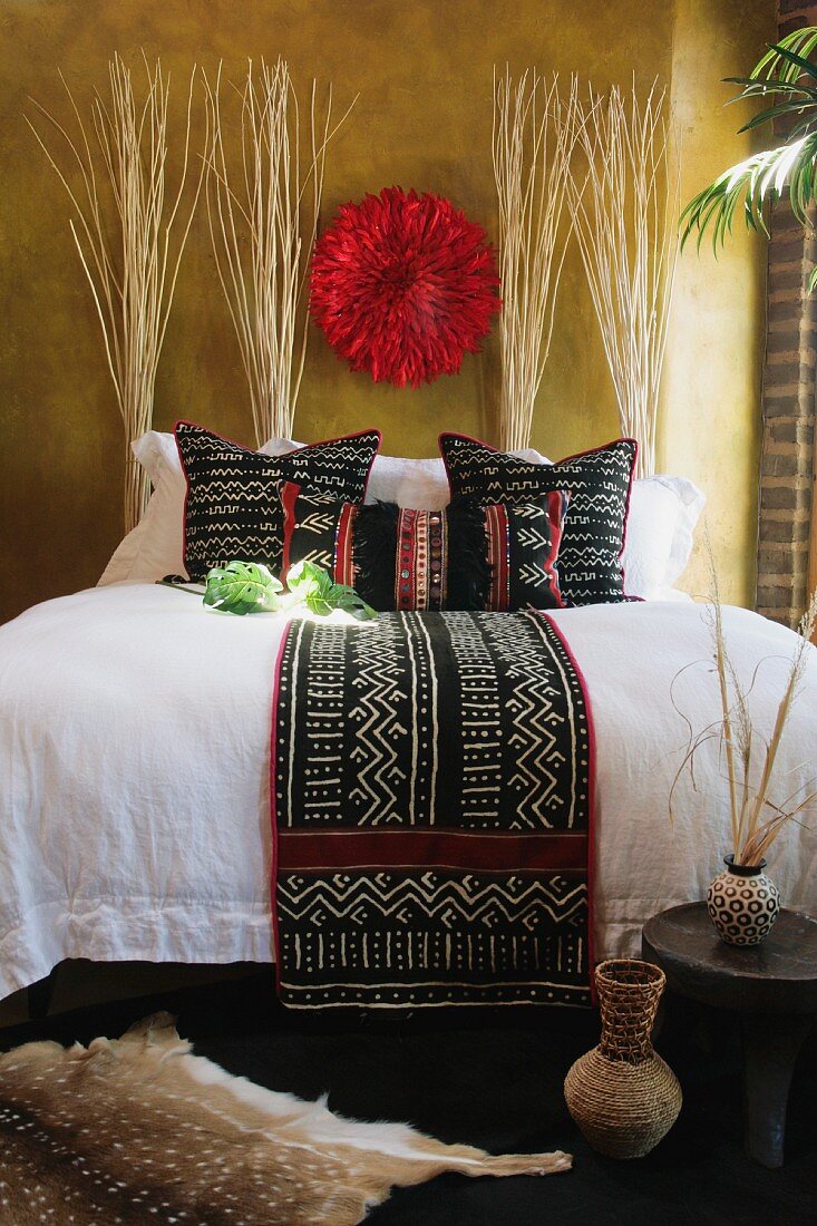 Doppelbett mit dunkler Tagesdecke in afrikanischem Stil auf weisser Bettwäsche und Bastschmuck an getönter Wand
