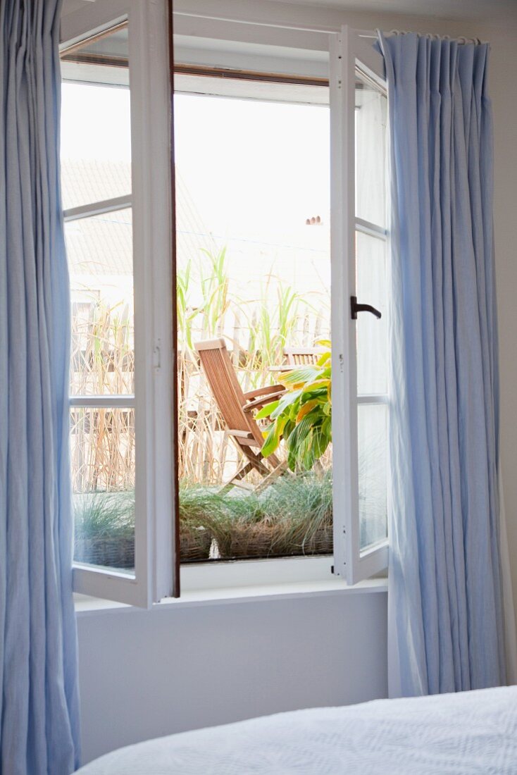 Offenes Fenster mit hellblauen Vorhängen im Schlafzimmer und Blick auf Terrasse