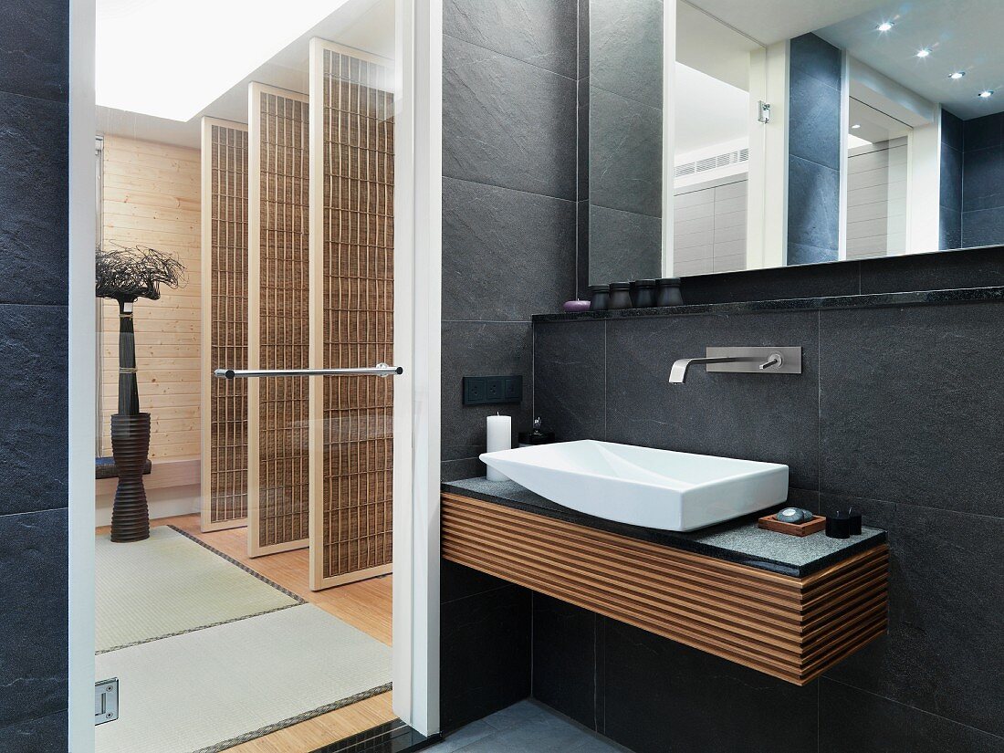 Designer Bad mit Waschtisch an grauen Wandfliesen neben Glastür und Blick in Vorraum mit raumhohen Paravent Türen