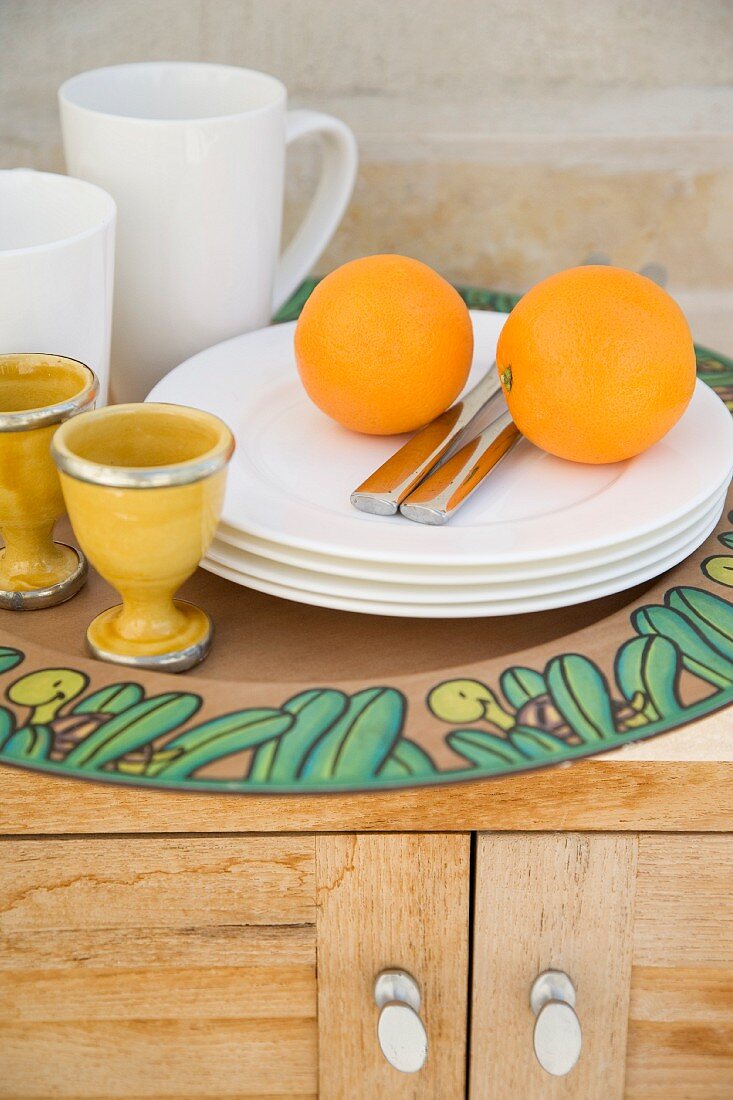 Oranges, stack of white plates, egg cups and mugs on tray on wooden sideboard