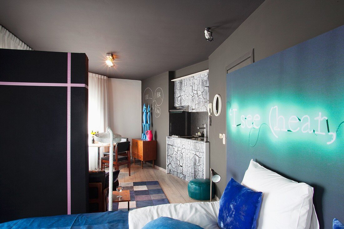 Bett an Wand mit montierter Leuchtschrift in offenem Schlafbereich und Blick auf Kochnische neben Essplatz