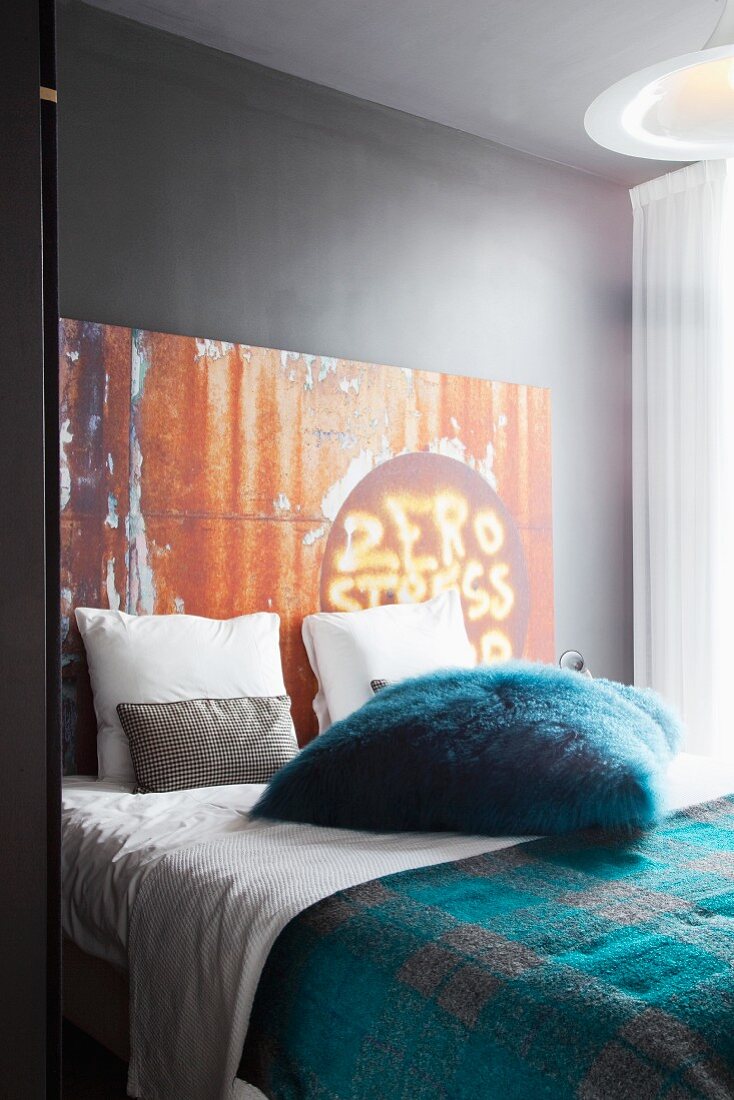 Scatter cushion with petrol blue faux fur cover on double bed against grey-painted wall with metal, rust-effect panel