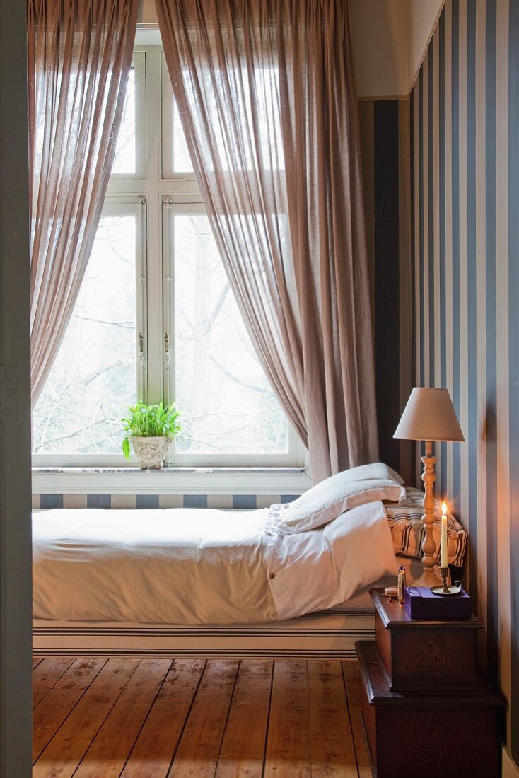 Schlichtes Bett neben Fenster mit gerafftem Vorhang in ländlichem Schlafzimmer mit Streifentapete