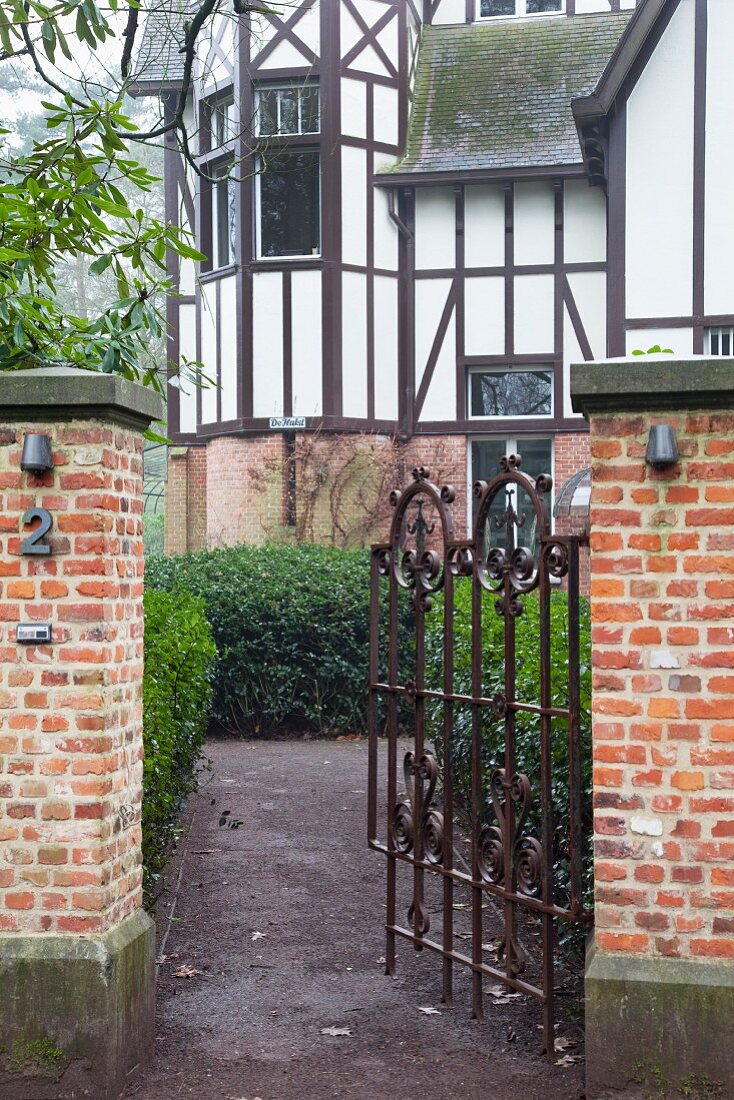 Open wrought iron garden gate with view into garden and traditional country house with half-timbered facade