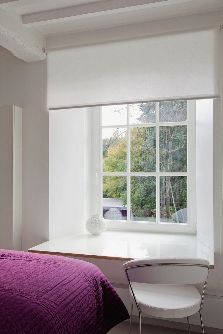 Weißes Springrollo über einer als Schreibtisch vergrößerten weißen Fensterbank und Designerstuhl; Ausschnitt violetter Tagesdecke im Vordergrund