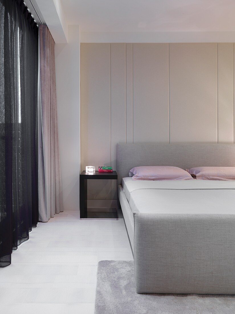 Modern bedroom with grey-upholstered double bed next to window with dark, transparent curtain