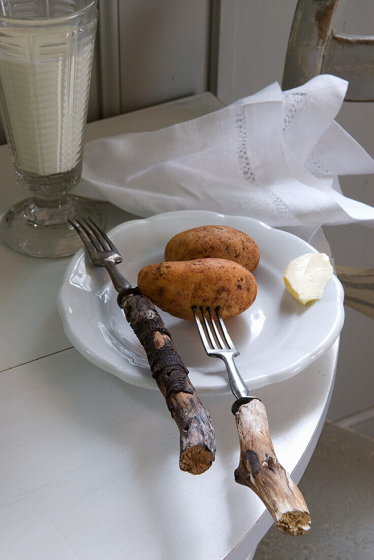 Rustikales Tischgedeck mit Kartoffeln auf weißem Teller, Altholz-Gabeln und Milch im Glas