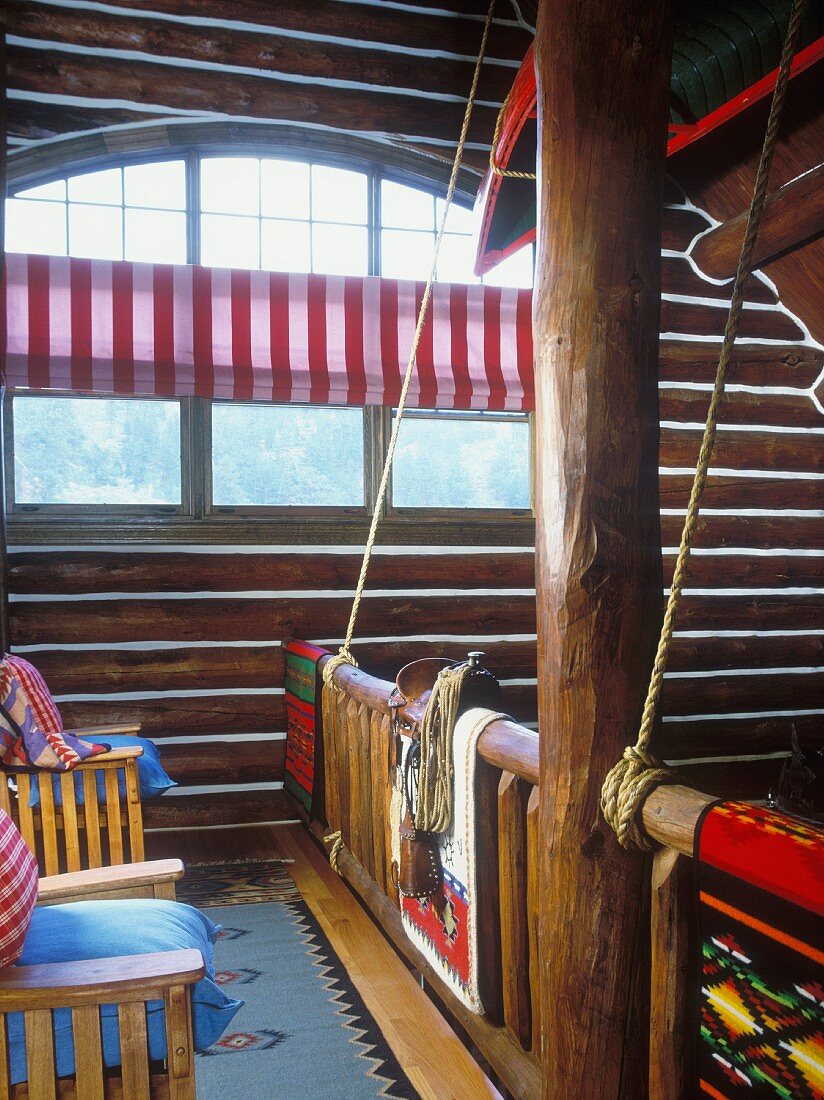 Blick von der Galerie eines Blockhauses auf ein halbrundes, großes Fenster, indianische Webteppiche und einem Kanu an der Decke