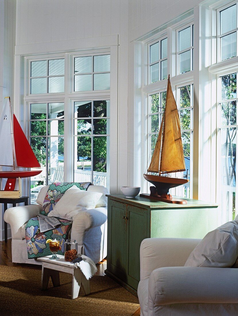 Maritimes Wohnzimmer mit Modell-Segelbooten, weissen Sesseln und einer großen Fensterfront
