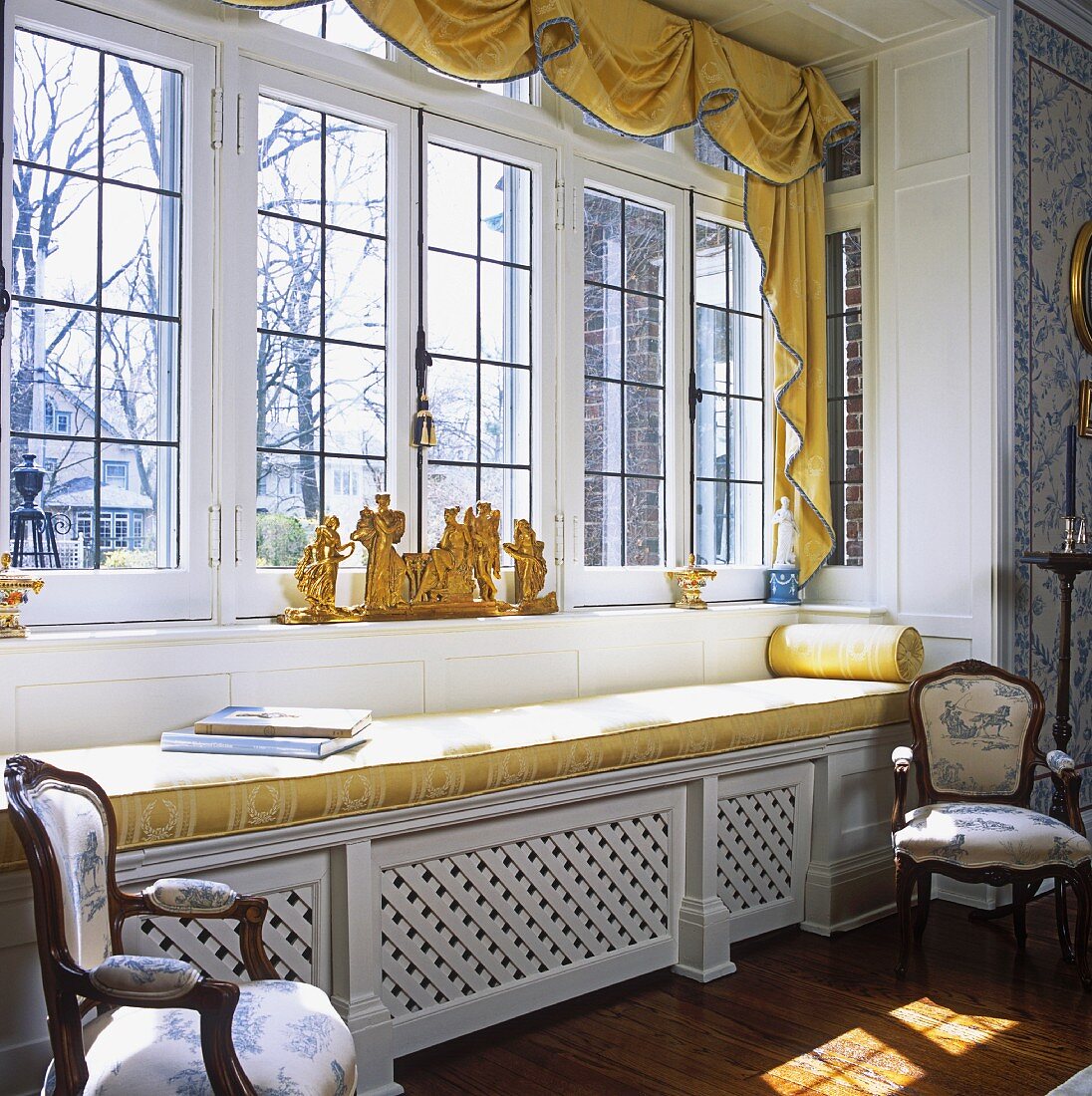 Fensterfront in einem Landhaus mit goldgelb gepolsteter Sitzbank und vergoldeter Skulptur