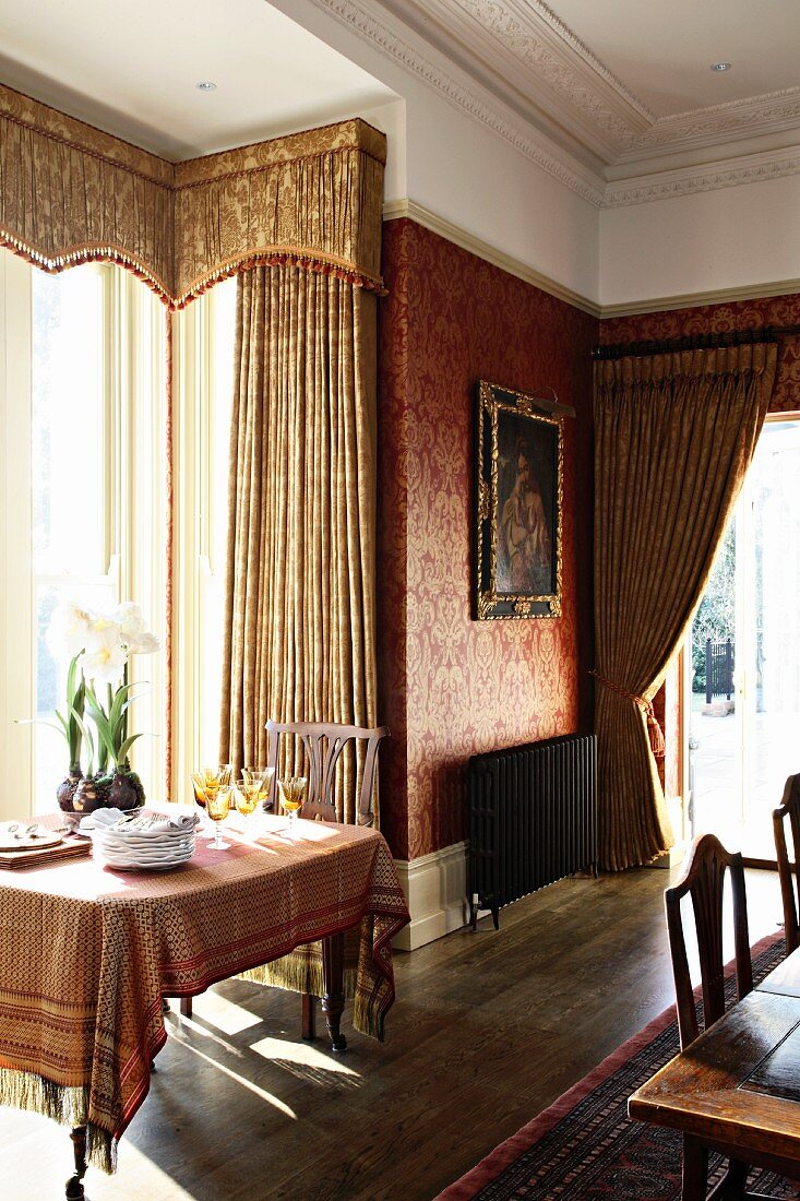 Gedeckter Tisch vor bodenlangen Brokatvorhängen und Schabracken an Fenster im Erker eines traditionellen Esszimmers