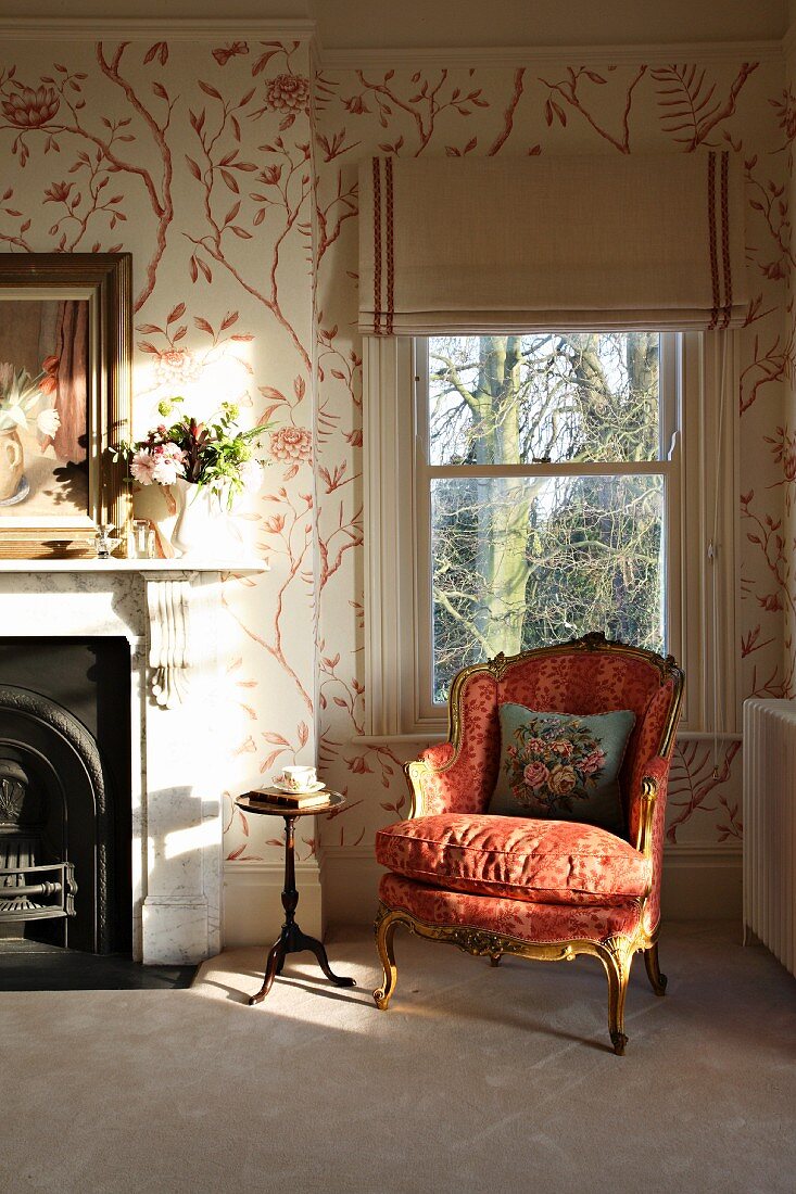 Rococo armchair in front of window and stencilled patterns on wall in traditional room with fireplace