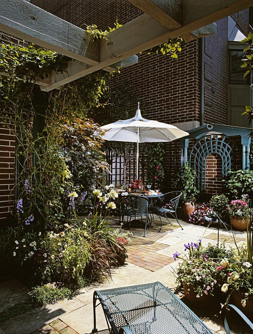 Bewachsene Gartenterrasse eines Backsteinhauses mit vielen Pflanzen, Blumentöpfen, einem Sonnenschirm, rundem Tisch und luftigen Stühlen