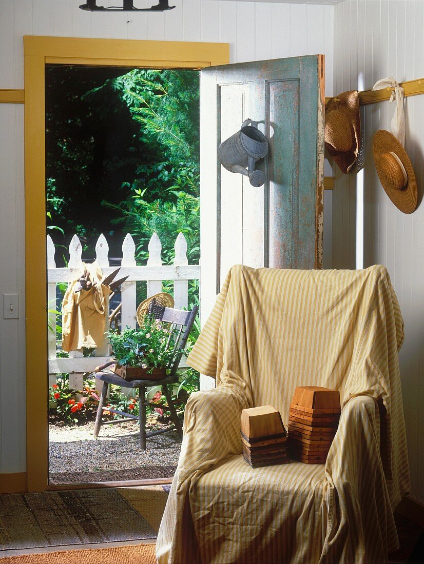 Ohrensessel mit gelb-weiss gestreiftem Überwurf, gestapelten Behältern darauf; dahinter eine offene Tür mit Blick auf einen weissen Gartenzaun und einem Blumentopf auf einem Stuhl