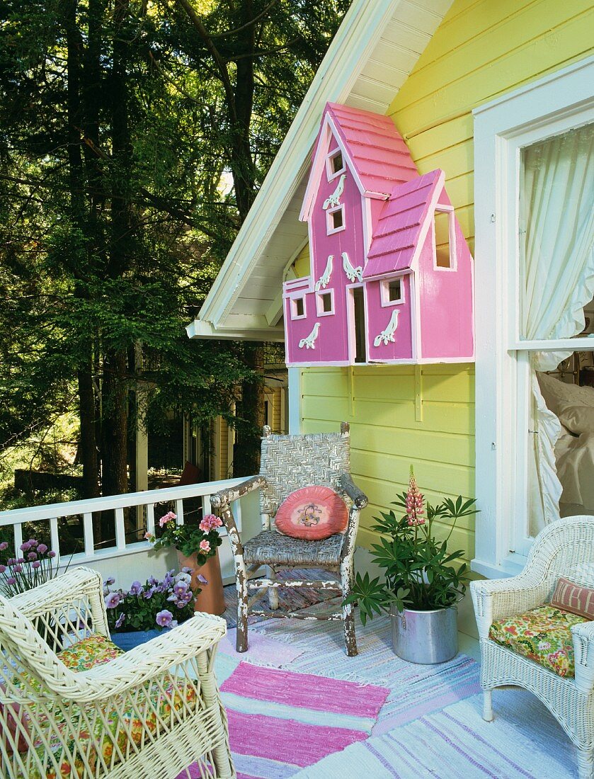 Terrasse mit einer Hausfassade in Gelb mit einem pinkfarbenem Vogelhäuschen, rosa-weiss gestreiftem Teppich und einem rustikalen Holzstuhl in der Ecke