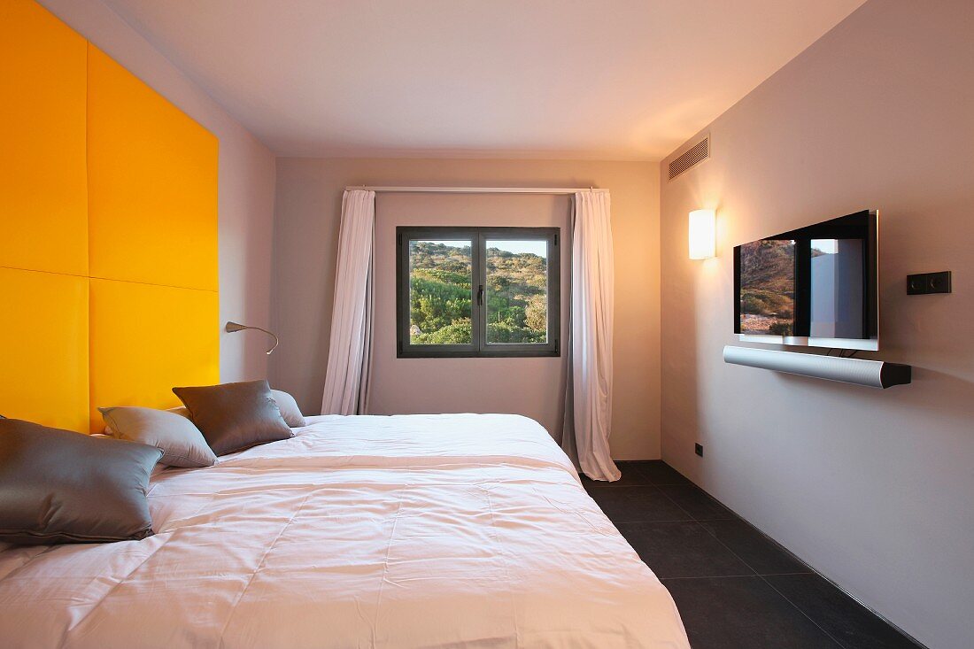 Moderner Schlafraum mit Bett vor gelbem gepolsterten Panel und an Wand befestigter Fernseher