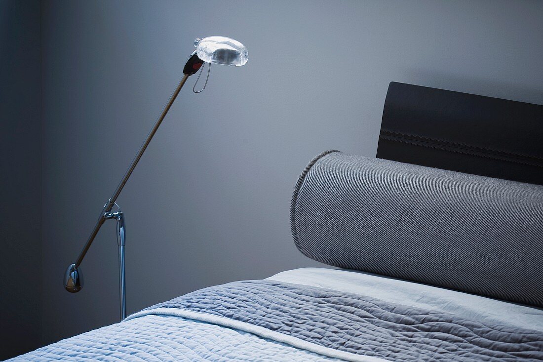 Moderne, kleine Stehlampe neben Bett mit Nackenrolle und Tagesdecke in Blaugrau-Tönen