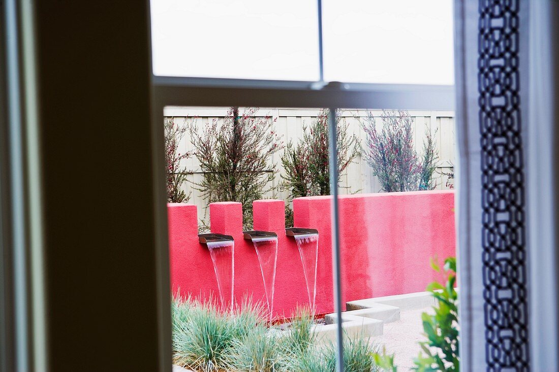 Blick durch ein Fenster auf rote Terrassenwand mit modernem Wasserfallbrunnen