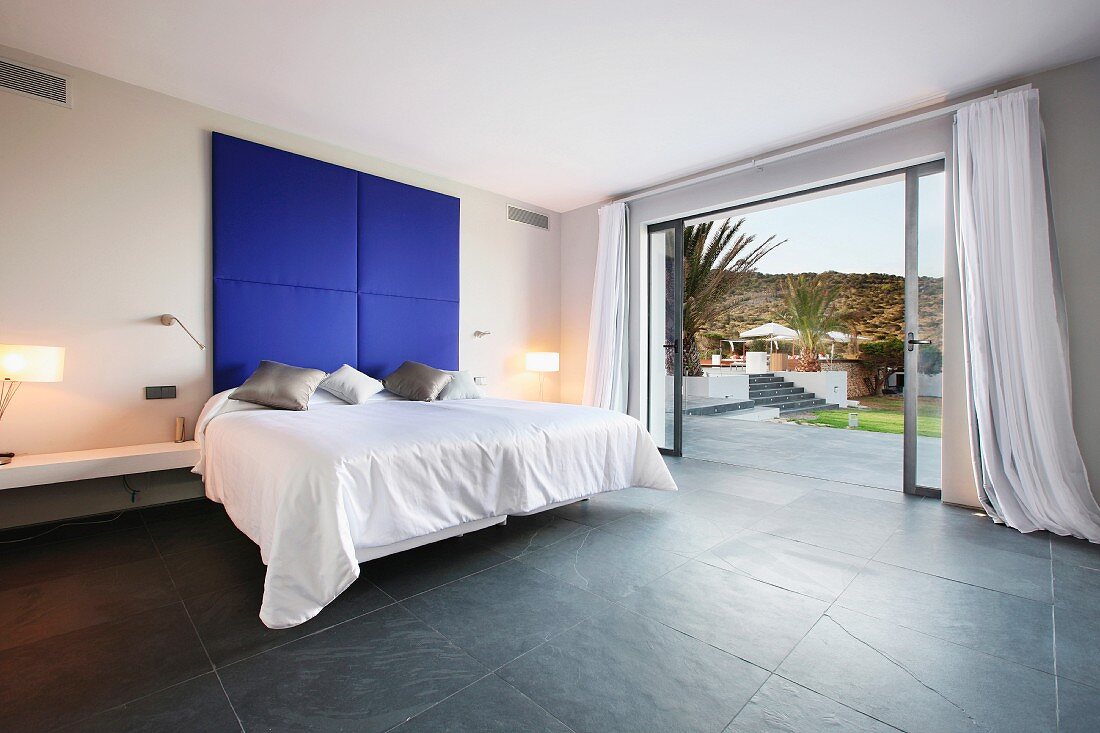 Bett vor blauer gepolstertem Panel an Wand in zeitgenössischem Schlafzimmer und Blick durch offene Glasschiebetür in Garten
