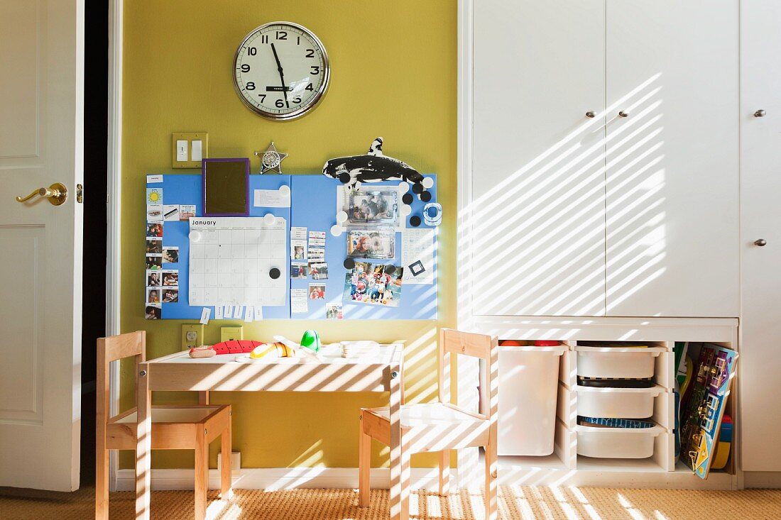 Kinderzimmer mit Tisch und Stühlchen vor Pinwand und Bahnhofsuhr an gelb getönter Wand