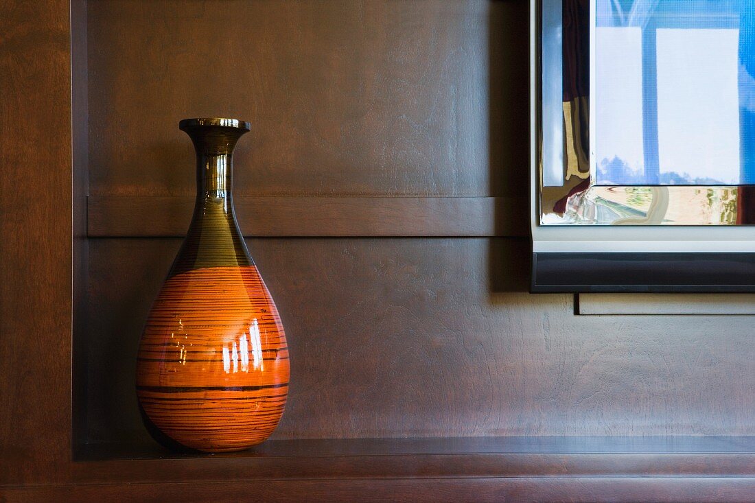 Zweifarbige, bauchige Vase und spiegelnder Rahmen in holzverkleideter Nische