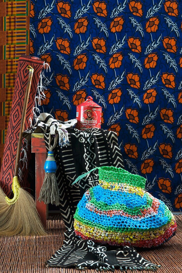 Gehäkelte Tasche aus bunten Streifen vor Hocker und geblümter Wandbehang in afrikanischem Stil